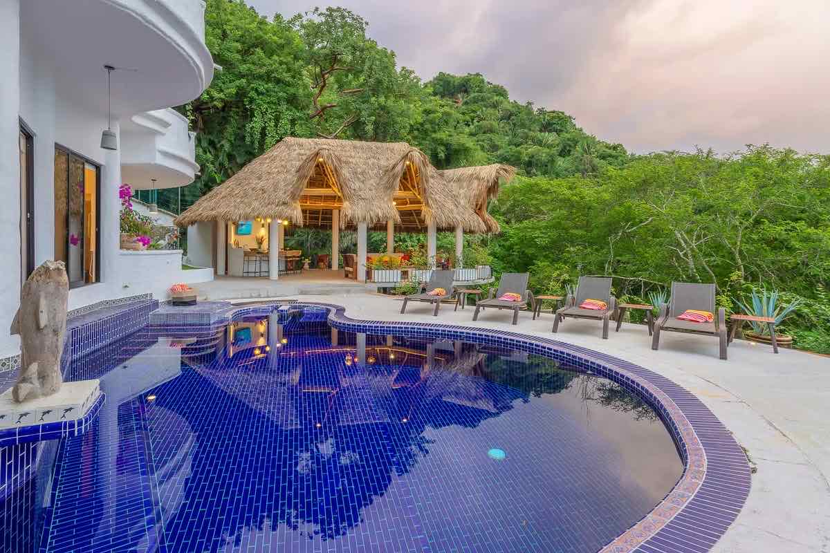 Alberca de azulejo azul junto a una palapa al anochecer, rodeada de selva